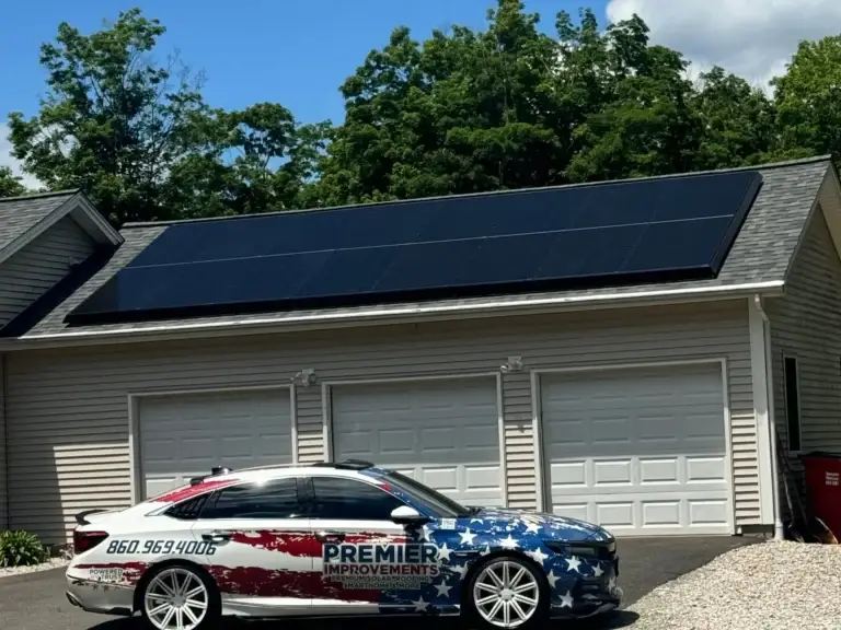 Ground-level view of a completed garage solar panel installation by Premier Improvements Solar in Connecticut, featuring sleek all-black panels on a shingled roof. A branded Premier Improvements Solar vehicle with patriotic red, white, and blue graphics is parked in front, highlighting the company’s local and veteran-owned pride.